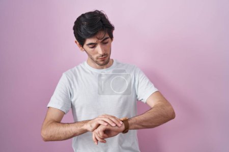Téléchargez les photos : Jeune homme hispanique debout sur fond rose vérifier l'heure sur la montre-bracelet, détendu et confiant - en image libre de droit