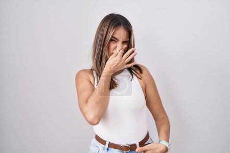 Téléchargez les photos : Jeune femme hispanique debout sur fond blanc sentant quelque chose de puant et dégoûtant, odeur intolérable, retenant souffle avec les doigts sur le nez. mauvaise odeur - en image libre de droit