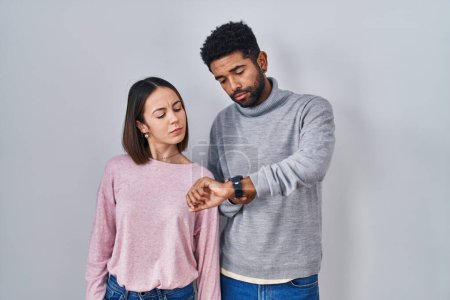 Téléchargez les photos : Jeune couple hispanique debout ensemble vérifier l'heure sur la montre-bracelet, détendu et confiant - en image libre de droit