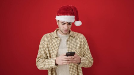 Foto de Joven hombre hispano con sombrero de Navidad usando teléfono inteligente sobre fondo rojo aislado - Imagen libre de derechos