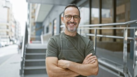 Photo for Cheerful middle age man with grey hair, full of joy and confidence, standing on a city street, arms crossed, flashing a handsome smile - Royalty Free Image