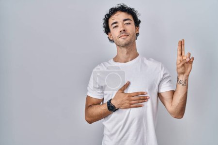 Téléchargez les photos : Homme hispanique debout sur un fond isolé souriant jurer avec la main sur la poitrine et les doigts vers le haut, faisant un serment de fidélité promesse - en image libre de droit