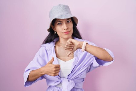 Photo for Young hispanic woman standing over pink background wearing hat doing thumbs up and down, disagreement and agreement expression. crazy conflict - Royalty Free Image