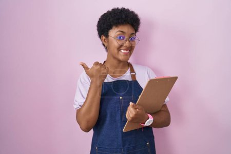Foto de Joven mujer afroamericana vistiendo delantal de camarera profesional sosteniendo portapapeles apuntando a la espalda con la mano y los pulgares hacia arriba, sonriendo confiado - Imagen libre de derechos