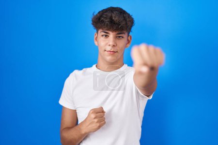 Foto de Adolescente hispana de pie sobre fondo azul golpeando puño para luchar, ataque agresivo y enojado, amenaza y violencia - Imagen libre de derechos
