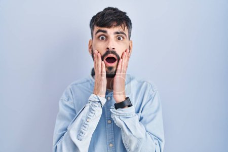 Photo for Young hispanic man with beard standing over blue background afraid and shocked, surprise and amazed expression with hands on face - Royalty Free Image