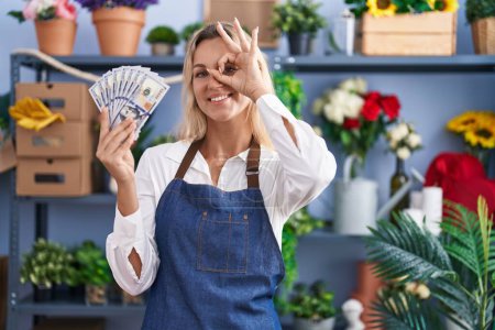 Téléchargez les photos : Jeune femme blonde travaillant à la boutique de fleuriste tenant des dollars souriant heureux faire ok signe avec la main sur les yeux en regardant à travers les doigts - en image libre de droit