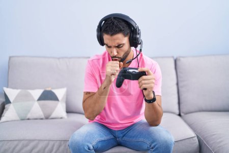 Photo for Hispanic young man playing video game holding controller sitting on the sofa feeling unwell and coughing as symptom for cold or bronchitis. health care concept. - Royalty Free Image
