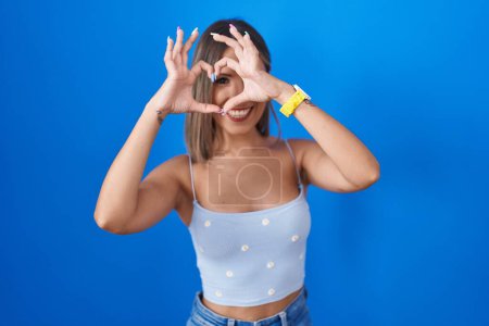 Photo for Young woman standing over blue background doing heart shape with hand and fingers smiling looking through sign - Royalty Free Image