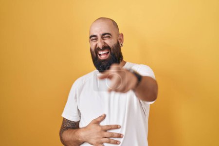 Téléchargez les photos : Jeune homme hispanique avec barbe et tatouages debout sur fond jaune se moquant de vous, pointant du doigt la caméra avec la main sur le corps, expression de honte - en image libre de droit