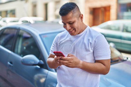 Téléchargez les photos : Jeune homme latin en utilisant smartphone appuyé sur la voiture à la rue - en image libre de droit