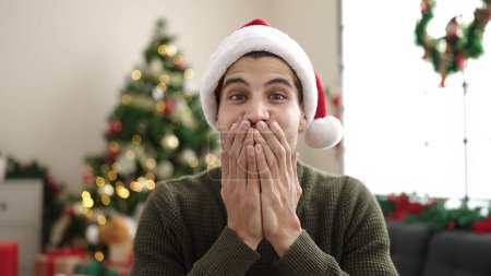 Téléchargez les photos : Jeune homme hispanique assis sur le canapé près de l'arbre de Noël avec expression surprise à la maison - en image libre de droit