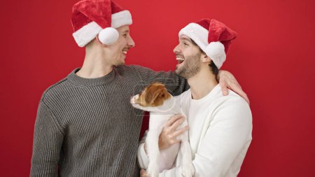 Téléchargez les photos : Deux hommes couple portant chapeau de Noël étreignant chien sur fond rouge isolé - en image libre de droit