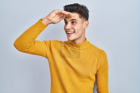 Photo for Young hispanic man standing over blue background very happy and smiling looking far away with hand over head. searching concept. - Royalty Free Image