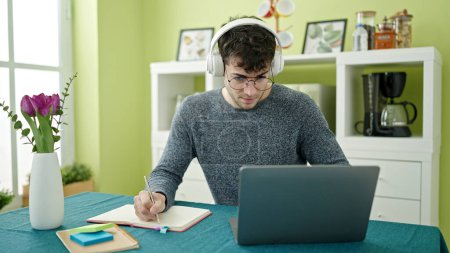 Téléchargez les photos : Jeune homme hispanique étudiant utilisant un ordinateur portable prenant des notes portant des écouteurs à la salle à manger - en image libre de droit