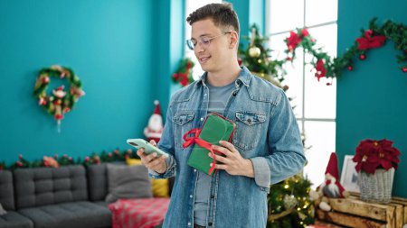 Foto de Joven hombre hispano usando smartphone sosteniendo regalo de Navidad en casa - Imagen libre de derechos