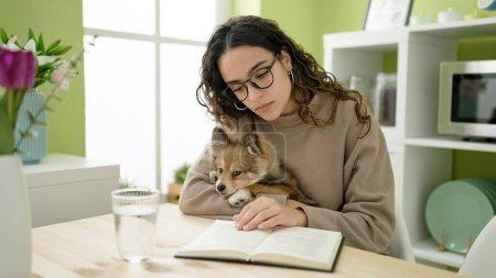 Photo for Young hispanic woman with dog sitting on table reading book at dinning room - Royalty Free Image