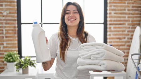 Photo for Young beautiful hispanic woman smiling confident holding folded towels and detergent bottle at laundry room - Royalty Free Image