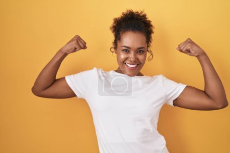 Photo for Young hispanic woman with curly hair standing over yellow background showing arms muscles smiling proud. fitness concept. - Royalty Free Image