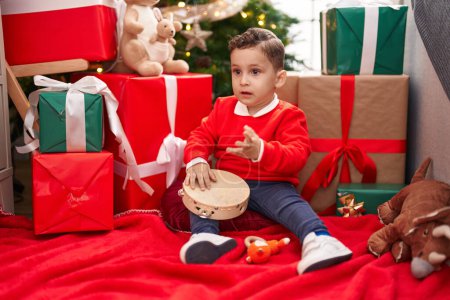 Photo for Adorable hispanic toddler playing tambourine sitting on floor by christmas gifts at home - Royalty Free Image