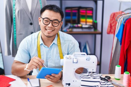 Téléchargez les photos : Jeune homme chinois tailleur souriant confiant en utilisant le pavé tactile au magasin de tailleur - en image libre de droit