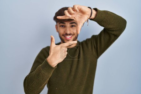 Téléchargez les photos : Homme arabe avec barbe debout sur fond bleu souriant faisant cadre avec les mains et les doigts avec le visage heureux. concept de créativité et photographie. - en image libre de droit