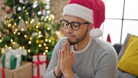 Téléchargez les photos : Homme hispanique priant assis près de l'arbre de Noël à la maison - en image libre de droit