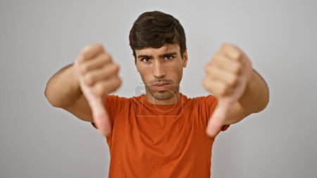 Photo for Cool portrait of a young hispanic guy making thumbs down gesture, a sign of negative lifestyle, failure, or bad fashion over isolated white wall background - Royalty Free Image