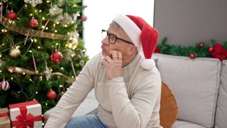 Téléchargez les photos : Homme aux cheveux gris du Moyen Âge assis sur un canapé près d'un sapin de Noël avec une expression ennuyeuse à la maison - en image libre de droit