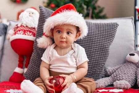 Photo for Adorable hispanic baby holding christmas decoration ball sitting on sofa at home - Royalty Free Image