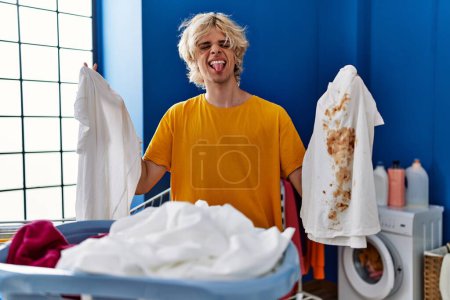 Foto de Joven sosteniendo camiseta blanca limpia y camiseta con mancha sucia sacando la lengua feliz con expresión divertida. - Imagen libre de derechos