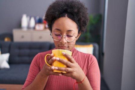 Téléchargez les photos : Femme afro-américaine sentant tasse de café assis sur le canapé à la maison - en image libre de droit