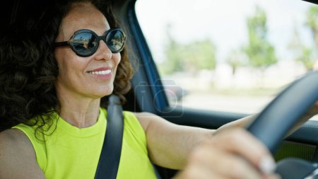 Téléchargez les photos : Moyen Age femme hispanique conduisant une voiture souriant portant des lunettes de soleil sur la route - en image libre de droit