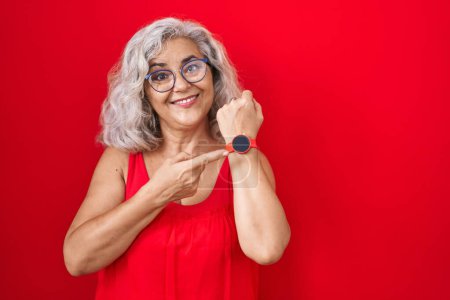 Foto de Mujer de mediana edad con el pelo gris de pie sobre el fondo rojo en la prisa que apunta a ver el tiempo, la impaciencia, mirando a la cámara con expresión relajada - Imagen libre de derechos