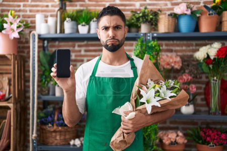 Téléchargez les photos : Jeune homme hispanique travaillant à la boutique de fleuristes montrant écran smartphone joues gonflées avec drôle de visage. bouche gonflée d'air, attrapant l'air. - en image libre de droit