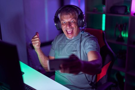 Téléchargez les photos : Homme hispanique aux cheveux gris jouant à des jeux vidéo avec smartphone pointant le pouce vers le côté souriant heureux avec la bouche ouverte - en image libre de droit
