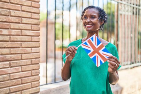 Téléchargez les photos : Moyen Age femme afro-américaine souriante confiante tenant drapeau du royaume uni dans la rue - en image libre de droit
