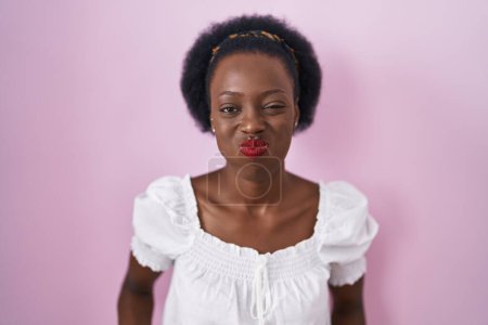 Foto de Mujer africana con el pelo rizado de pie sobre fondo rosa hinchando mejillas con cara divertida. boca hinchada de aire, expresión loca. - Imagen libre de derechos