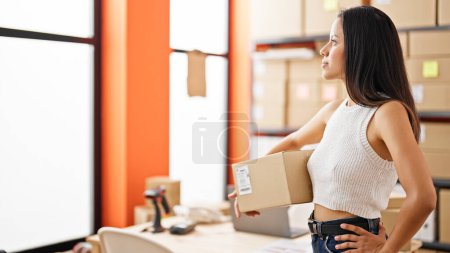 Téléchargez les photos : Jeune belle hispanique femme commerce électronique travailleur entreprise tenant paquet regardant jeter la fenêtre au bureau - en image libre de droit