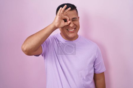 Photo for Young hispanic man standing over pink background doing ok gesture with hand smiling, eye looking through fingers with happy face. - Royalty Free Image