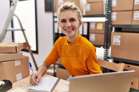 Téléchargez les photos : Jeune femme blonde ecommerce travailleur d'affaires en utilisant un ordinateur portable prenant des notes au bureau - en image libre de droit