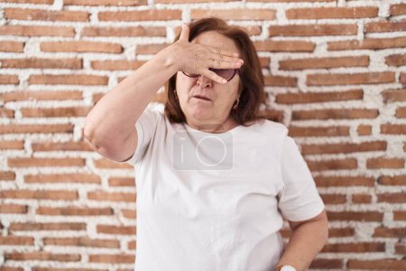 Téléchargez les photos : Femme âgée avec des lunettes debout sur le mur de briques couvrant les yeux avec la main, l'air grave et triste. concept de non-visibilité, de dissimulation et de rejet - en image libre de droit
