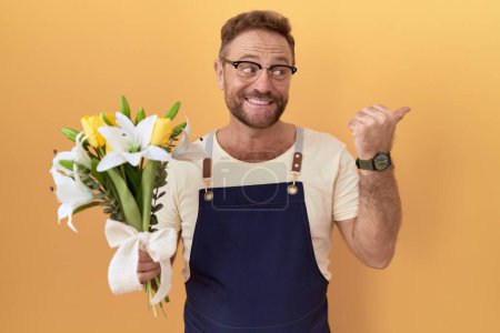 Téléchargez les photos : Moyen Age homme avec barbe fleuriste magasin tenant des fleurs souriant avec le visage heureux regardant et pointant vers le côté avec le pouce vers le haut. - en image libre de droit
