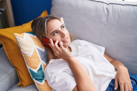 Téléchargez les photos : Jeune femme blonde parlant sur le smartphone couché sur le canapé à la maison - en image libre de droit