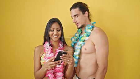 Téléchargez les photos : Couple touriste homme et femme portant le lei hawaïen à l'aide d'un smartphone sur fond jaune isolé - en image libre de droit