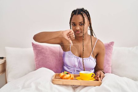 Téléchargez les photos : Femme afro-américaine avec des tresses tenant un plateau avec de la nourriture pour le petit déjeuner dans le lit avec un visage en colère, signe négatif montrant aversion avec les pouces vers le bas, concept de rejet - en image libre de droit