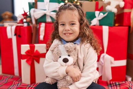 Photo for Adorable blonde toddler hugging teddy bear sitting on floor by christmas tree at home - Royalty Free Image