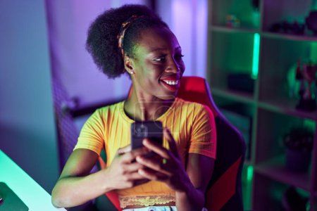 Téléchargez les photos : Femme afro-américaine streamer souriant confiant en utilisant smartphone à la salle de jeux - en image libre de droit