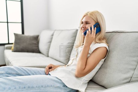 Téléchargez les photos : Jeune femme blonde parlant au téléphone assis sur le canapé à la maison - en image libre de droit