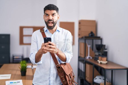 Foto de Hombre hispano con barba usando teléfono inteligente en la oficina enojado y loco gritando frustrado y furioso, gritando con ira mirando hacia arriba. - Imagen libre de derechos